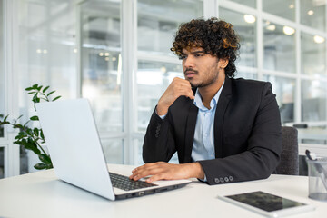 Focused young man in business attire working at his laptop in a brightly lit modern office. His...
