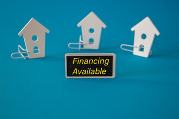 Financing available symbol. Wooden houses sits next to a wooden black board with the words Financing available. Beautiful blue background. Business and federal housing administration concept