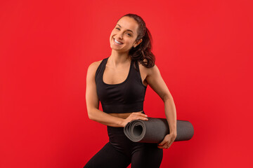 A woman standing while holding a yoga mat and smiling brightly. She appears joyful and ready for her yoga practice.