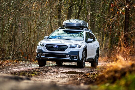 White Subaru Outback car with ski box drives on a muddy road in the forest. Large SUV with all-wheel drive on a broken road.