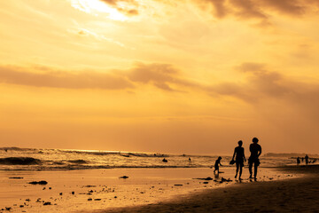 Two adults walking and a child playing along the beach in Canggu, Bali Indonesia at sunset, with...
