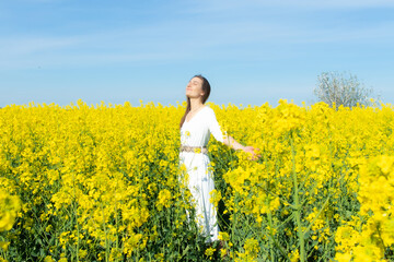 Young beautiful woman in a white dress close up in the middle of the yellow field with the radish flowers closeup on the blue sky background. Republic of Belarus, Brest region