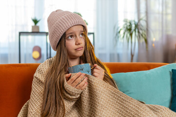 Sick ill girl child wrapped in blanket shivering from cold drinking hot herbal medical tea in unheated home apartment. Unhealthy Caucasian teenager try to warming up sitting on sofa in living room.
