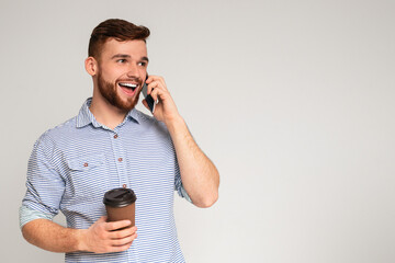 Handsome millennial guy talking on cellphone and holding coffee to go on studio background, panorama