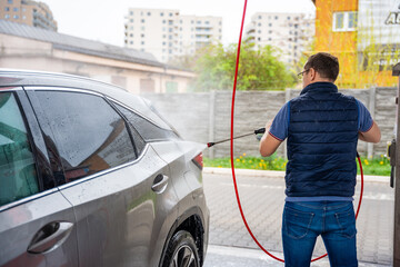 Young man washes his car at a self-service car wash using a hose with pressurized water and foam.