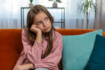 Portrait of calm brunette little girl sitting alone on couch in living room at home. Preteen teenage kid thinking dreaming imagining leaning head on hand. Pose of boredom disinterest fatigue concept. - obrazy, fototapety, plakaty