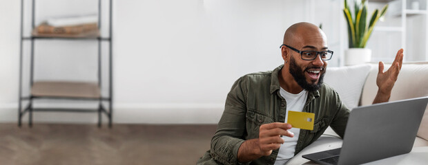 African American man showing excitement while holding a card and using a laptop, ample copy space...