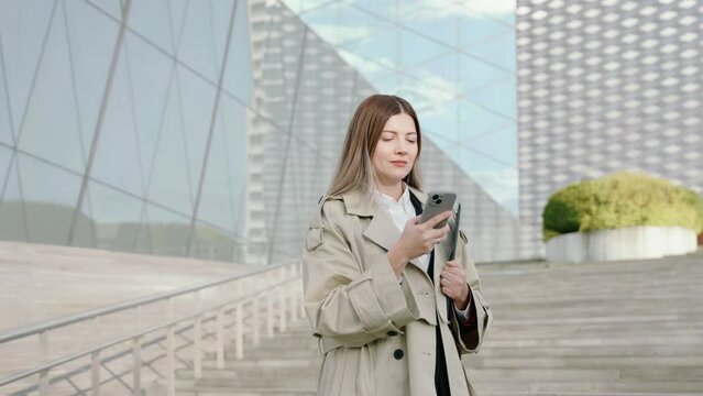 Young businesswoman in a white shirt and coat with a folder in her hand talking on the phone in front of office buildings