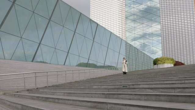 Young businesswoman in a white shirt and coat with a folder in her hand talking on the phone in front of office buildings