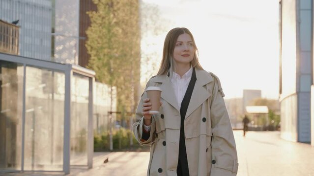 Stylish young woman in a coat walking during sunset around the city holding a cup of coffee 