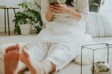 A woman wearing loosing white clothes playing cellphone on the carpet, leisure at home