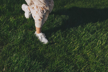 Female legs in summer dress and sneakers. Feet in white sneakers in green grass (grass, shoes, freedom). Summertime concept. Rest in park. Colorful and positive mood. 