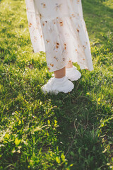 Female legs in summer dress and sneakers. Feet in white sneakers in green grass (grass, shoes, freedom). Summertime concept. Rest in park. Colorful and positive mood. 