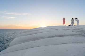 un groupe de jeunes filles de dos sur un rocher blancs face à un coucher de soleil sur la mer