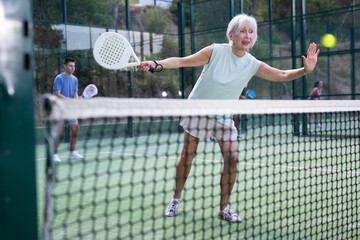 Focused aged woman playing friendly paddleball match on outdoor summer court. Senior people sports...