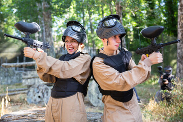 Tactical young man and woman standing back-to-back, aiming and shooting with paintball guns during...