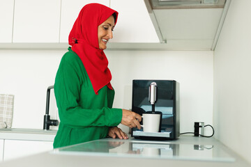 A woman wearing a red and green hijab is standing in a kitchen, pouring hot water into a coffee mug. She appears focused as she prepares her morning drink.