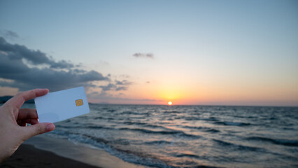 A woman's hand holds a white bank card against a sunset backdrop on a beach. Concept of money...