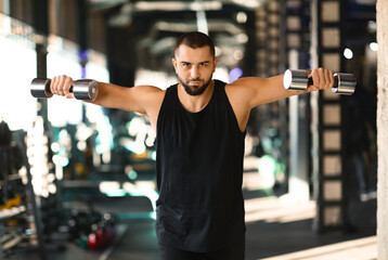 A man is standing while holding two dumbbells in his hands. He appears to be focusing on his...