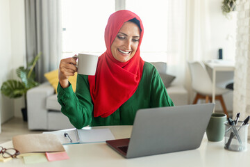Muslim woman is seated at a table, working on her laptop and sipping coffee from a cup. She appears...