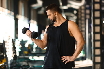 A focused, athletic man with a beard is standing inside a well-equipped gym, wearing a sleeveless...