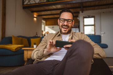 Adult man with eyeglasses play video games on his phone at home