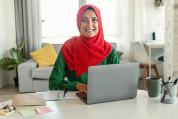 Muslim woman is seated at a table, engrossed in her laptop. The screen casts a soft glow over her...