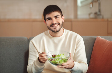 A man is seen sitting on a couch, holding a bowl of salad in his hands. He appears to be enjoying...