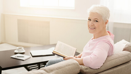 Elegant senior lady relaxing with favorite book at home, free space