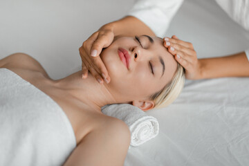 Fototapeta na wymiar A woman lies on a spa bed while a professional gently massages her face, focusing on pressure points and areas of tension, top view