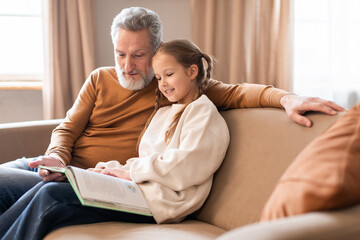 Senior man and a little girl are sitting on a couch, engrossed in reading a book together....