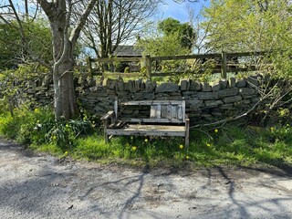 A weathered wooden bench sits in front of a dry stone wall with verdant foliage, nestled in a quiet roadside spot on, Upper Allerton Lane, Bradford, UK