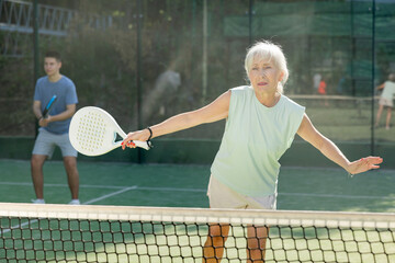 Senior woman in shorts playing padel tennis on court. Racket sport training outdoors.