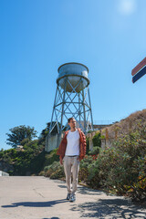 Person walking by Alcatraz Island water tower, dressed casually in white top, light pants, and...