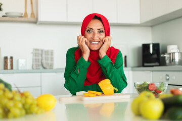 A woman wearing a red and green hijab is seated at a kitchen table, with various kitchen utensils...