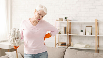 Senior woman is diligently cleaning a living room floor with a mop. She is focused on removing dirt...