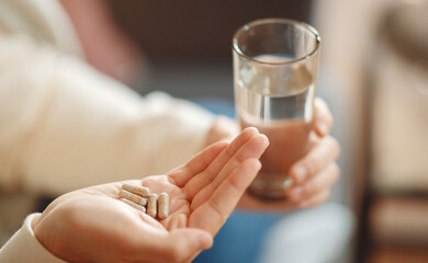A close-up view illustrates man holding several pills in one hand and clear glass of water in the...
