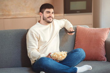 A young man lounges comfortably on a grey sofa, holding a bowl of popcorn on his lap and a remote...