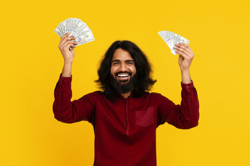 Indian man with long hair and a beard is standing while holding up two stacks of money. The money...