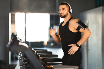 A focused young man with a beard, wearing a black tank top and headphones, stands beside a...