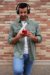 Vertical photo of a handsome charming man isolated in a brick wall, using smartphone to listen music wearing headphones.
