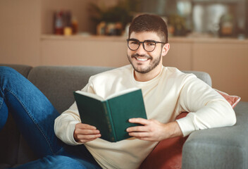 A man wearing eyeglasses is seated on a couch, engrossed in a book in his hands. The room appears...