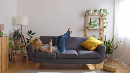Full angle view of happy young caucasian woman using mobile phone while lying a couch at home.