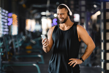 A man in a sleeveless top and headphones smiling as he selects a playlist on his smartphone in gym....