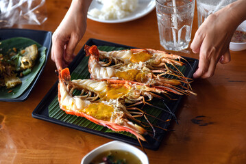 A close-up photo of a hand supporting a plate of large grilled shrimp that had been cut in half,...
