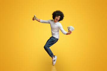 Young and rich. Happy african american guy dancing on tiptoes with money fans, orange studio...