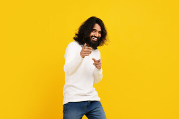 Indian man with long hair and a beard is standing in front of a bright yellow background, pointing...