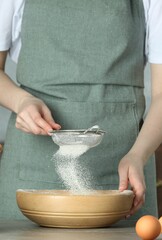 Woman sieving flour into bowl at table in kitchen, closeup