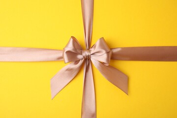 Beige satin ribbon with bow on yellow background, top view