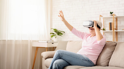 Excited senior woman enjoying cyber world, using VR glasses at home, free space